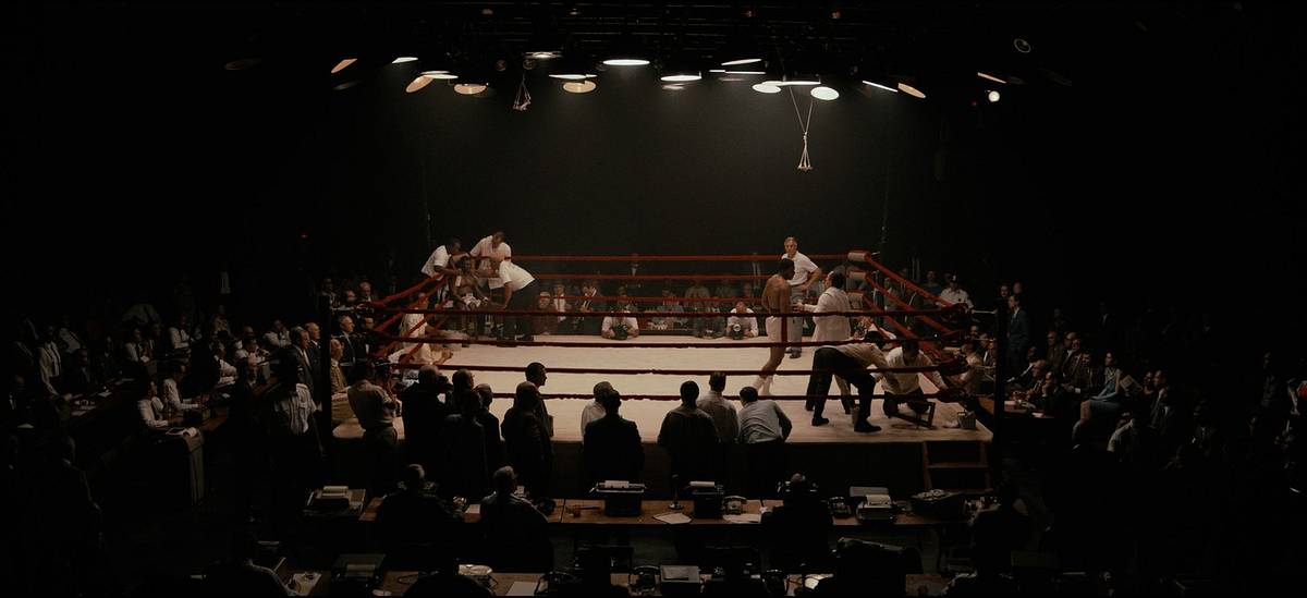 A film still looking down on a dimly lit arena, with a boxing ring surrounded by spectators and ringside press tables.