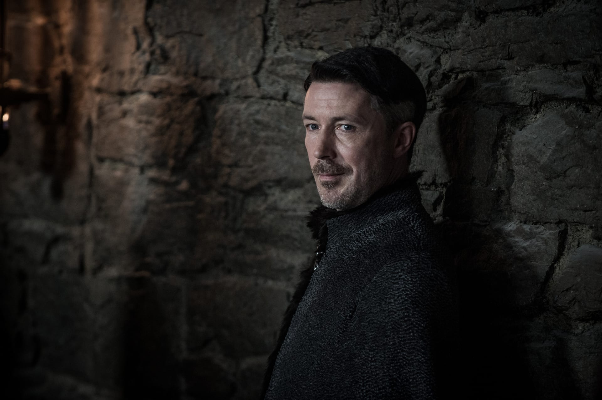 Littlefinger gives a sneaky look as he leans against the stone walls of Winterfell.