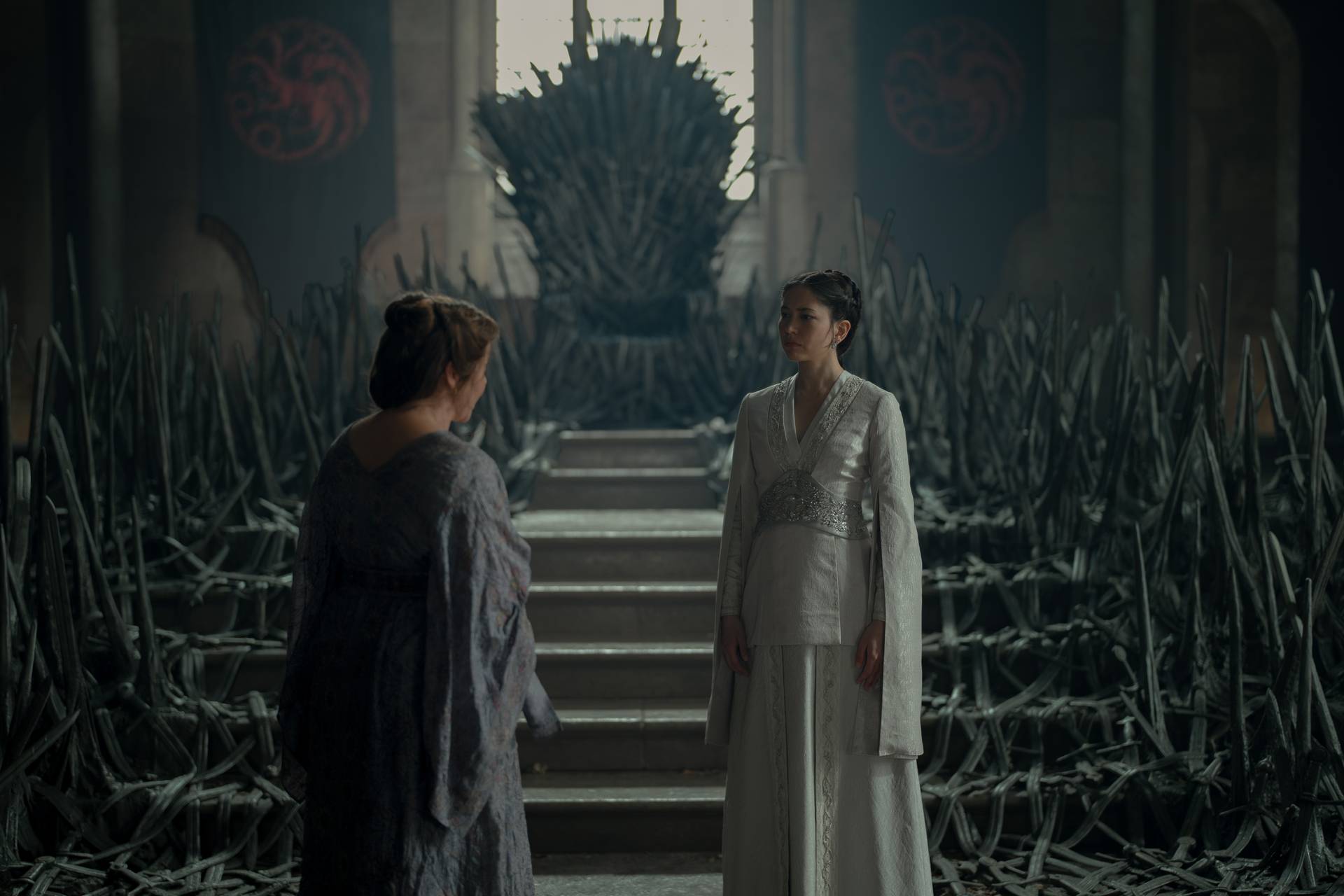 A wide shot from a dramatic television production set inside a dimly lit, stone throne room. Two women stand in the foreground facing one another in conversation. On the right, a young woman with dark hair styled in an updo wears a structured, flowing cream-colored gown with long split sleeves and an intricate wide belt. On the left, an older woman with her brown hair pinned up is seen from behind, dressed in a loose-fitting lavender or grey patterned robe. Between them in the background, stone steps lead up to a massive, imposing throne constructed from twisted black swords, which towers toward a large window. Dozens of melted, jagged swords fan out across the floor on either side of the steps. In the upper background, two dark banners bearing a red, multi-headed dragon sigil hang from the stone walls.
