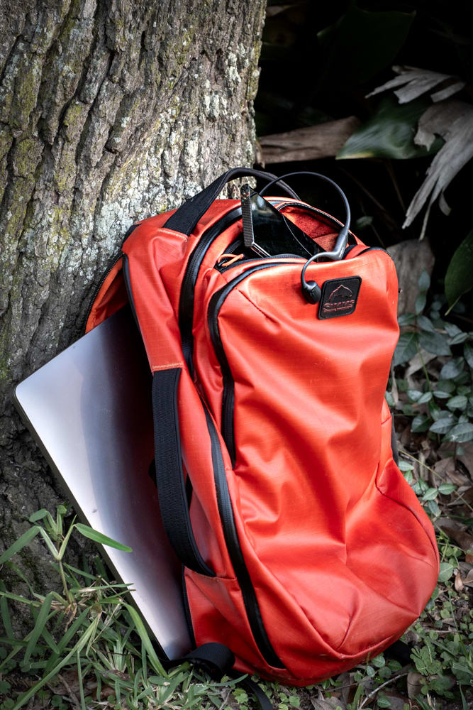 The Simms Tailwind backpack leaning against a tree, with a laptop partially in the side pocket.