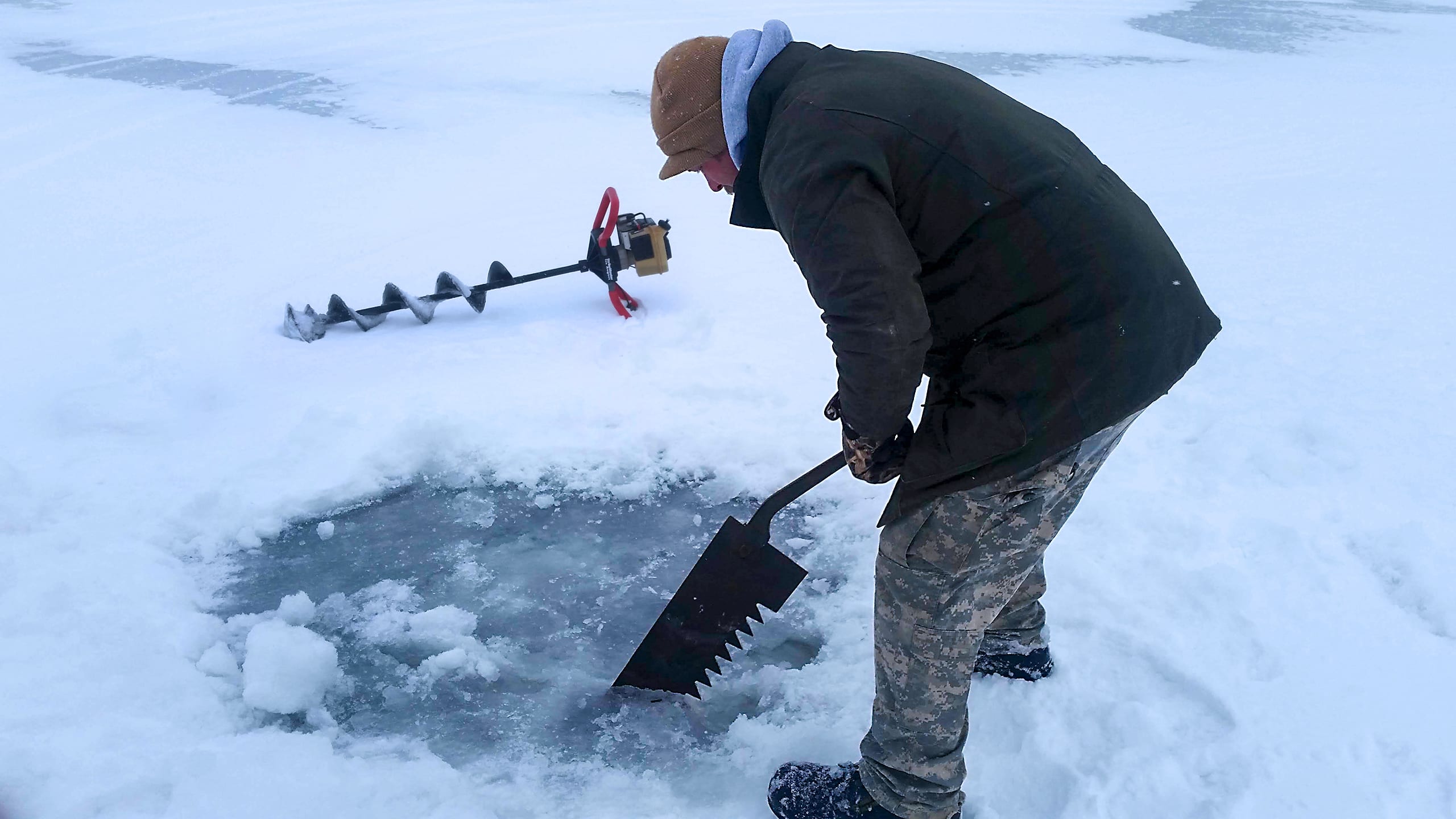 Angler cutting an ice hole with an auger and saw for pike spearing.