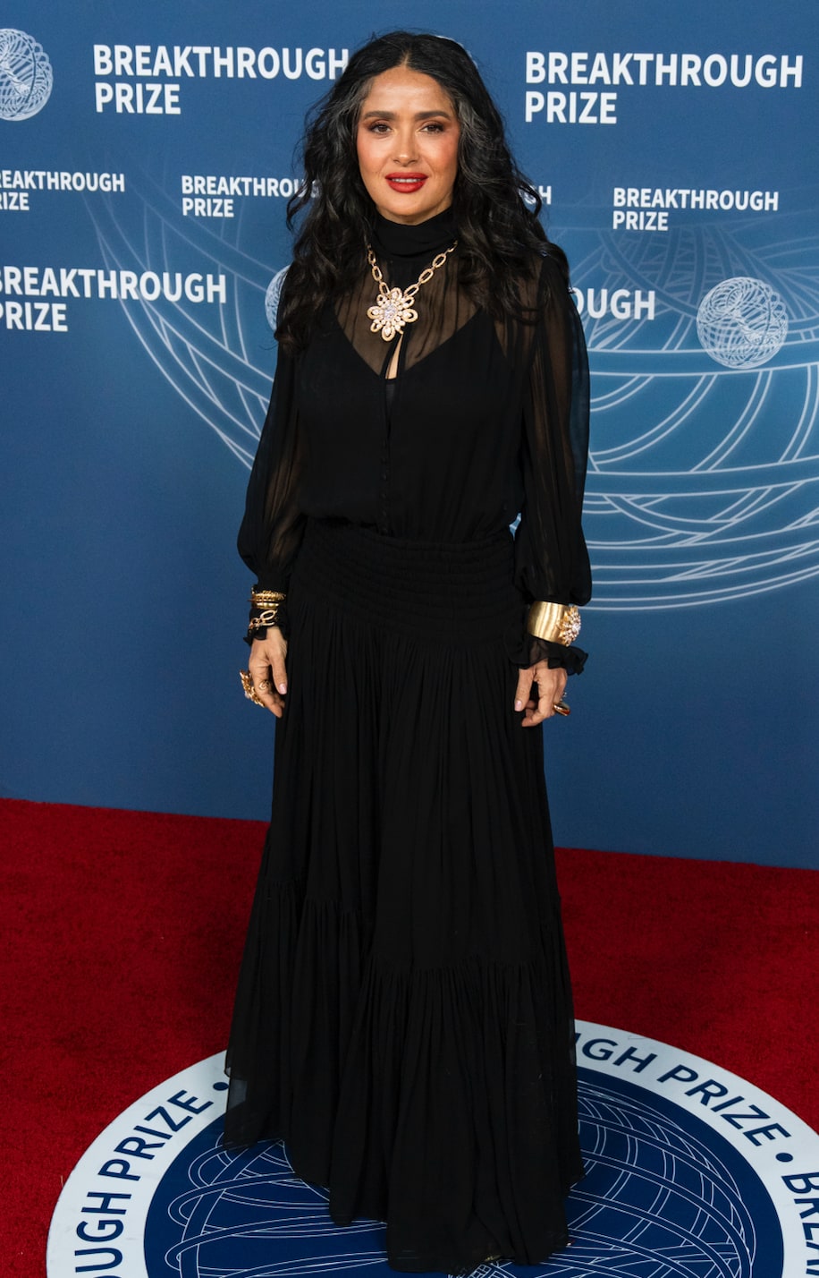 Salma Hayek poses on the red carpet in an all-black maxi dress from YSL and gold jewelry.