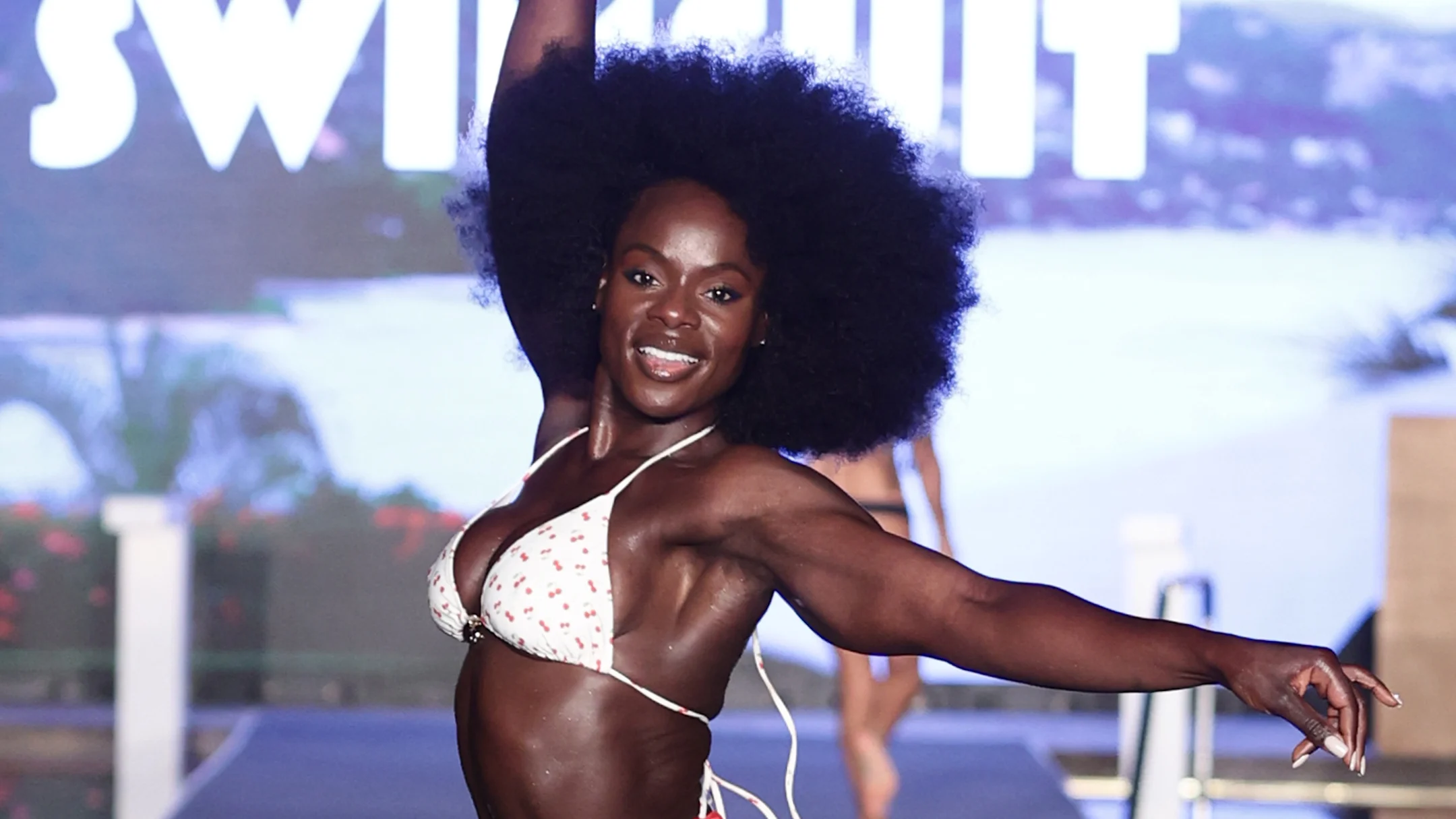 Tunde Oyeneyin in a cherry print string bikini at the SI Swimsuit 2025 runway show, arms wide open and beaming