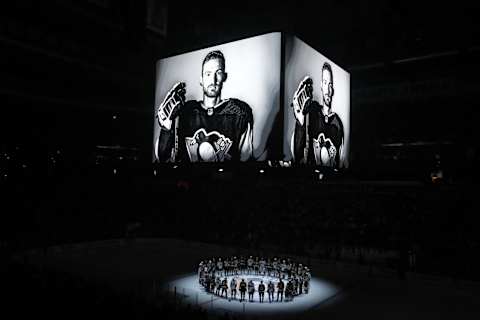 Oct 30, 2023; Pittsburgh, Pennsylvania, USA;  Members of the Anaheim Ducks and the Pittsburgh Penguins stand at center ice for a moment of silence at the PPG Paints Arena to honor former Penguin forward Adam Johnson (on scoreboard) who was tragically killed in a hockey related accident in Europe. Mandatory Credit: Charles LeClaire-USA TODAY Sports