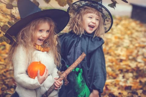 Two adorable blond little girls dressed up like witches for Halloween and grinning, one with a pumpkin