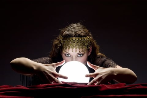 A female fortune-teller with gold headdress and her arms raised near a glowing crystal ball