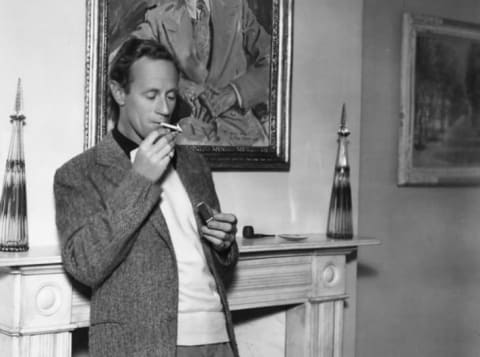 British actor Leslie Howard lighting a cigarette by the fireplace at his home in 1938.