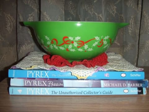 A green Pyrex mixing bowl with red ribbons and holly on it, sitting on top of three pyrex collecting books.