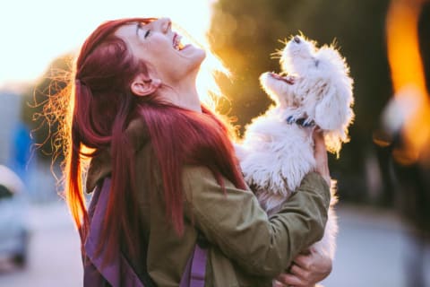 A red haired woman holding a white dog, both laughing.