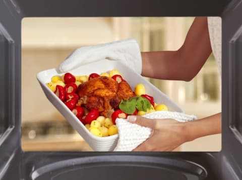 A woman holds up a baking dish with meat and potatoes in front of a microwave.