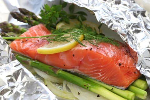 Raw salmon on top of asparagus spears and garnished with dill and lemon, all in a bed of aluminum foil.