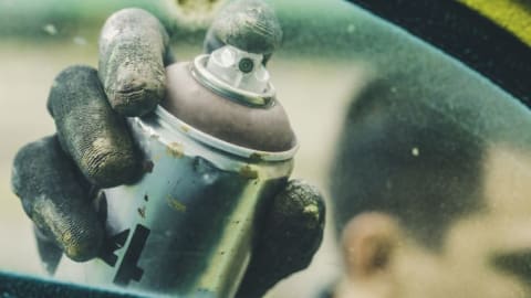 A gloved hand holding a spray paint can with a finger on the trigger.