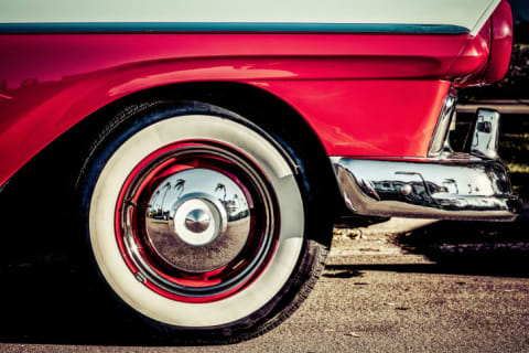 A hubcap of an old-fashioned red car with whitewall tires that show palm trees reflected in the hubcaps