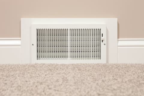 A white HVAC register set into a tan wall with neutral-colored carpet in the foreground.