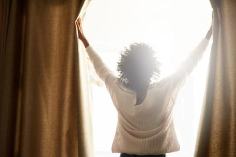 A woman in a white sweater with her back turned to us is opening heavy curtains, letting the sunlight stream in.