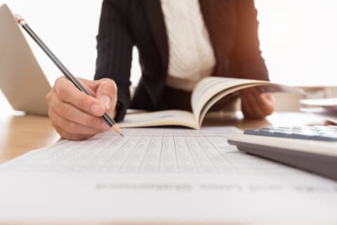 A person holding a pencil over a piece of paper with a book open in the background.