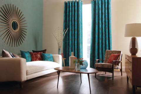 Heavy turquoise drapes adorn the windows in a modern-looking living room with a low white couch and sunburst mirror.