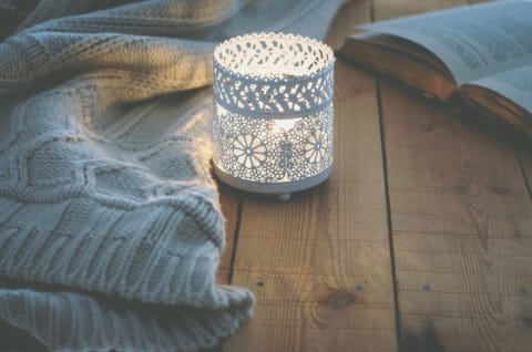 A candle in a white candle holder sitting on top of a rustic wood table. A knit blanket and an open book lie nearby.