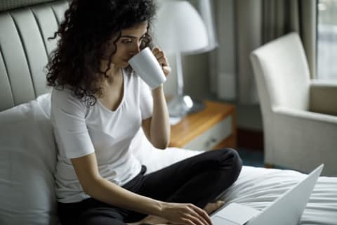 woman drinks from cup and uses laptop on hotel bed