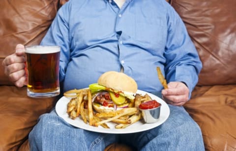 A man, shown from the neck down, sitting on a couch holding a beer with a burger and fries on a plate in his lap.