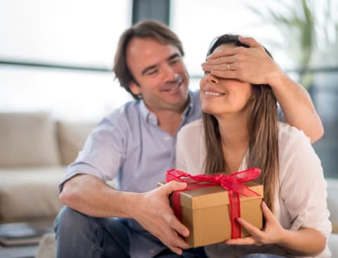 A man puts his hand over his partner's eyes as he hands her a gift.