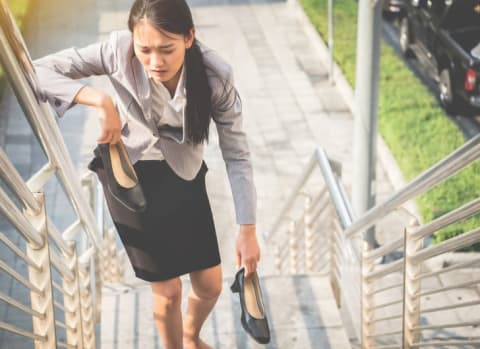 A woman walking up stairs, in pain, holding her black high heels.