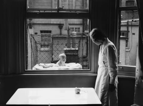 A nanny supervising a baby suspended in a wire cage attached to the outside of a high tenement block window.