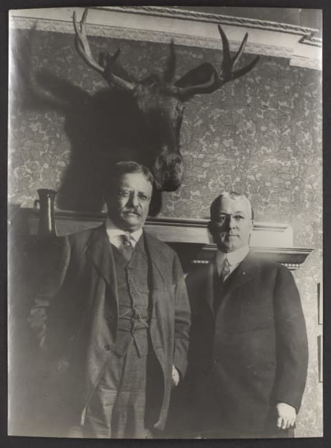 Theodore Roosevelt and professor Goode of Chicago University standing in front of a mounted moose head over professor Goode's fireplace.