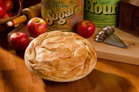 A picture of Apple Works's apple pie surrounded by apples and a container of sugar.