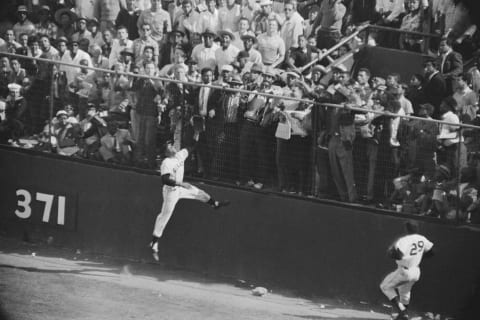 (Original Caption) Giant centerfielder Willie Mays scales the right centerfield wall in an attempt to snare drive by Philadelphia’s Harry Anderson in the ninth inning here. As the ball glances off the wall, right fielder Willie Kirkland (lower left), comes up and both watch the ball get away, as Anderson scores a double. Giants won, 1-0.