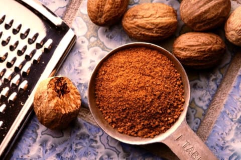 A tablespoon of grated nutmeg on a table surrounded by whole nutmegs and a grater.