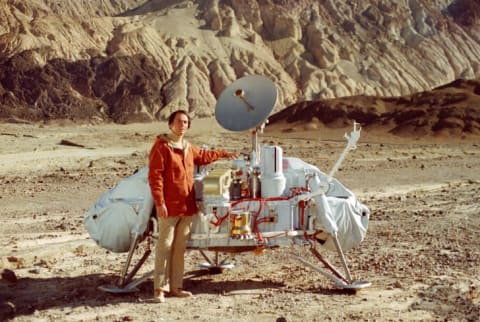 Carl Sagan standing with a model of the Viking Lander.